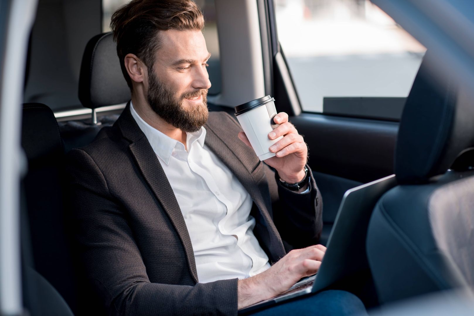 Ein bärtiger Mann sitzt auf dem Rücksitz eines Autos und arbeitet konzentriert an seinem Laptop.